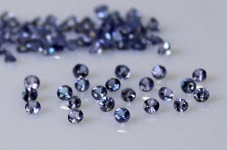 May include: A collection of small, round, blue gemstones. The gemstones are scattered on a white surface.