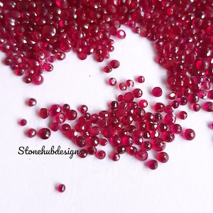 May include: A large quantity of small, red ruby gemstones. The gemstones are faceted and have a variety of sizes and shapes. The gemstones are scattered on a white surface.