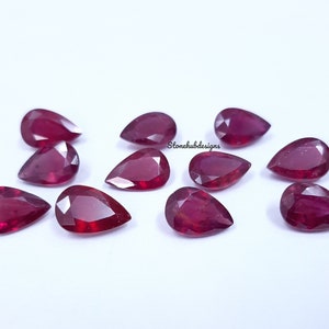 Pode incluir: Uma coleção de pedras preciosas de rubi facetadas em forma de pêra. As pedras são de uma cor vermelha profunda e rica e estão dispostas sobre uma superfície branca. As pedras preciosas são cortadas para maximizar a reflexão da luz e o brilho. A imagem é bem iluminada, destacando a clareza e a cor dos rubis.