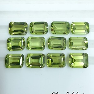 May include: Twelve green peridot gemstones, cut in a rectangular emerald shape, are arranged in three rows of four on a white surface. The gemstones are faceted and have a translucent appearance.