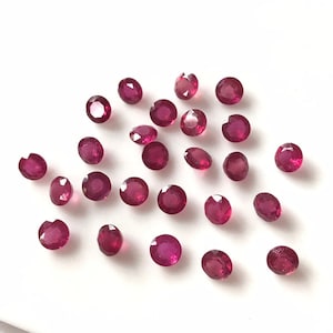 May include: A collection of 25 round, faceted ruby gemstones. The rubies are a deep red colour and are scattered on a white surface.