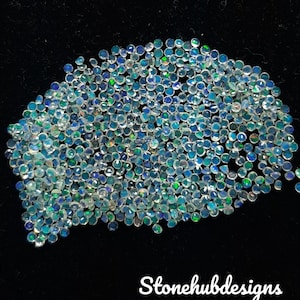 Puede incluir: Una colección de pequeñas gemas redondas y facetadas con un brillo iridiscente. Las gemas muestran una gama de colores, incluyendo azules y verdes. El fondo es negro, y el texto "Stonehubdesigns" es visible.