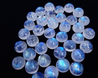 Puede incluir: Una colección de cabochones redondos de piedra lunar blanca con destellos azules iridiscentes. Las piedras son lisas y pulidas, y están dispuestas en un montón suelto.