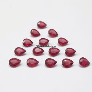 May include: A collection of 16 pear-shaped ruby gemstones, each with a deep red hue. The gemstones are arranged in a triangular pattern on a white surface.