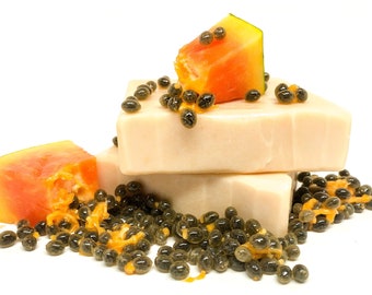 May include: Three bars of white soap with papaya fruit and seeds on top. The soap is stacked on top of each other. The papaya fruit is orange and yellow and the seeds are black.