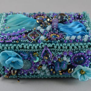 May include: A rectangular box covered in intricate beadwork featuring shades of turquoise, teal, and purple. The surface is embellished with small glass beads, faceted crystals, and fabric ribbon flowers in blue, creating a textured, layered appearance.