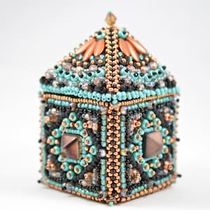 Puede incluir: Una pequeña caja de cuentas con un diseño geométrico intrincado. La caja está hecha con cuentas turquesa, negras, cobre y doradas. La parte superior de la caja tiene forma de pirámide.