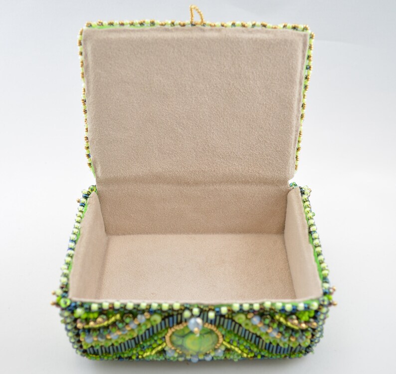 May include: A green beaded jewelry box with a light beige interior. The box is decorated with green, blue, and gold beads.