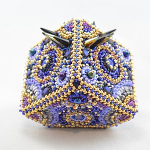 May include: A beaded sculpture in the shape of a stylized animal head. The sculpture is covered in small, colorful beads in shades of blue, purple, gold, and black. The head has two black, pointed horns.