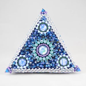 May include: A handcrafted, triangular beaded object in shades of blue, white, and purple. The pyramid-shaped piece features intricate beadwork with a central circular design and three additional circular accents. The edges are outlined in white beads.
