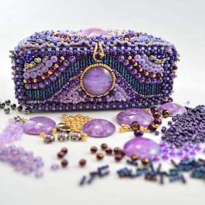 May include: A purple beaded box with a large purple cabochon in the center. The box is surrounded by loose beads in various shades of purple, gold, and silver.
