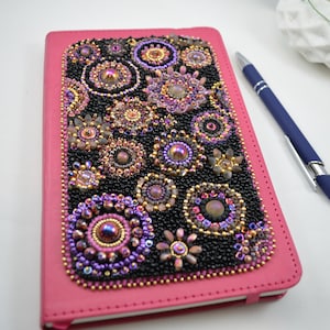 May include: A pink leather-bound journal with a black beaded cover featuring a floral design with iridescent beads.