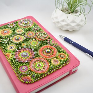 Puede incluir: Un diario de cuero rosa con una cubierta de cuentas que presenta un diseño floral colorido. Las cuentas son en tonos de verde, rosa, naranja y amarillo.