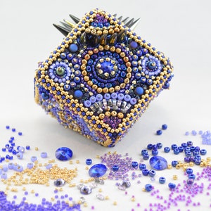 Puede incluir: Una caja cuadrada hecha a mano cubierta de intrincados abalorios. La caja presenta una combinación de cuentas azules, doradas y moradas, con una parte superior puntiaguda de color oscuro. Cuentas sueltas y cristales de colores similares están esparcidos.