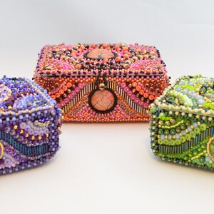 May include: Three small, intricately beaded boxes with different colored stones and designs. The boxes are purple, orange, and green.
