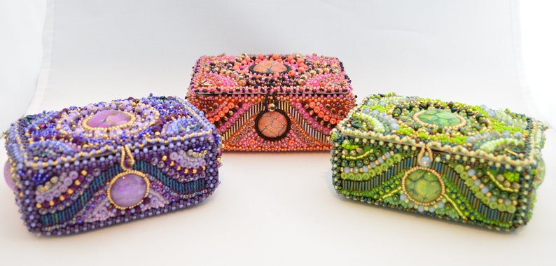 May include: Three small, intricately beaded boxes with different colored stones and designs. The boxes are purple, orange, and green.