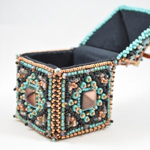 May include: A handcrafted, square beaded box with a hinged lid, open to reveal a black interior. The box is adorned with turquoise, black, and copper-colored beads, and features geometric patterns and square accents. The box is a decorative item.