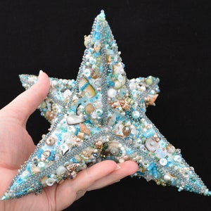 May include: A handcrafted, five-pointed star decoration. The star is covered in a mosaic of small, iridescent beads, shells, and sequins in shades of blue, white, and beige. The edges are outlined with a silver-colored chain. The overall effect is a coastal or nautical theme.