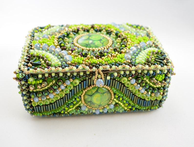 May include: A small, square box covered in green, blue, and gold beads. The box has a decorative, swirling pattern and a green and gold center piece.