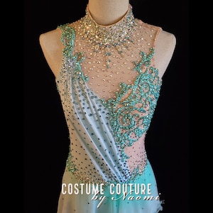 May include: A turquoise and white figure skating costume with a high neckline and a sheer bodice. The costume is embellished with rhinestones and a floral design. The text "Costume Couture by Naomi" is visible at the bottom of the image.