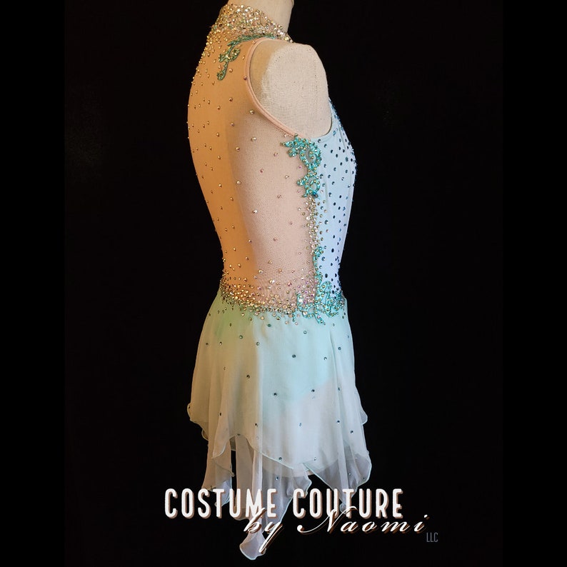 May include: A light blue and peach figure skating dress with a sheer bodice and a layered skirt. The dress is decorated with rhinestones and beads. The dress is on a white dress form. The dress form is on a black background. The text "COSTUME COUTURE by Naomi LLC" is at the bottom of the image.
