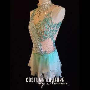May include: A light blue and white figure skating dress with a high neckline and a beaded design. The dress has a sheer bodice with a floral pattern and a layered skirt.