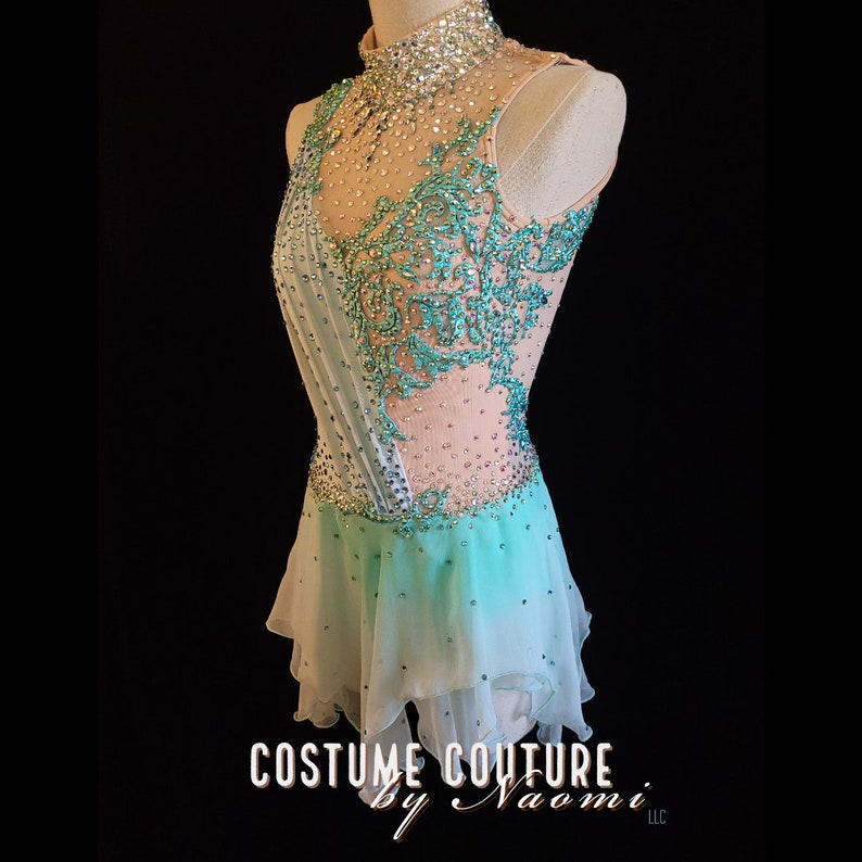 May include: A light blue and white figure skating dress with a high neckline and a beaded design. The dress has a sheer bodice with a floral pattern and a layered skirt.