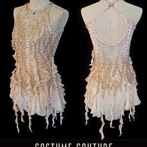 May include: A light pink figure skating dress with a halter neckline and a beaded bodice. The dress has a layered skirt with ruffled edges and a sheer overlay. The back of the dress has a keyhole opening and a criss-cross strap design. The dress is adorned with rhinestones and sequins.
