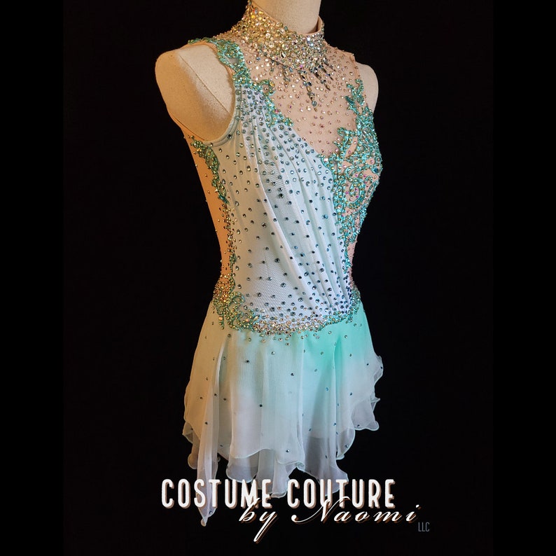 May include: A light blue and white figure skating dress with a high neck and a sheer bodice. The dress is embellished with rhinestones and sequins. The skirt is layered and has a scalloped hem. The dress is designed by Costume Couture by Naomi LLC.