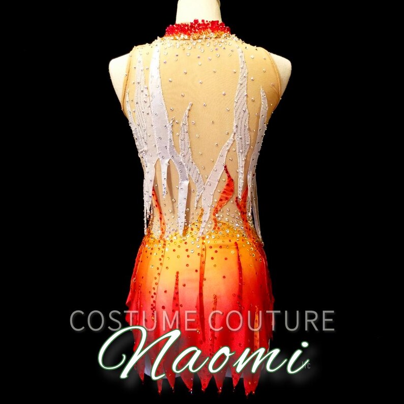 K&ouml;nnte beinhalten: Ein rot-orange-gelber Tanzanzug mit einem wei&szlig;en Flammenmuster und Strasssteinen. Der Tanzanzug befindet sich auf einem Mannequin und tr&auml;gt den Text "Costume Couture Naomi" am unteren Rand.