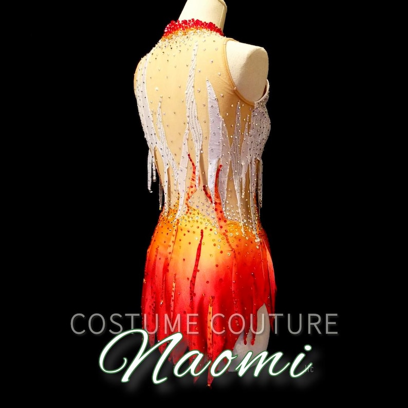 K&ouml;nnte beinhalten: Ein rot-orange-gelbes Trikot mit wei&szlig;en Fransen und Strasssteinen. Das Trikot ist so gestaltet, dass es wie Flammen aussieht. Das Trikot befindet sich auf einer Schaufensterpuppe. Der Text "Costume Couture Naomi" befindet sich am unteren Rand des Bildes.