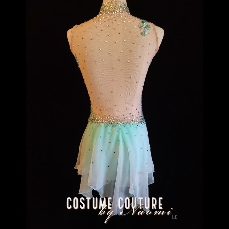 May include: A light blue and nude figure skating dress with a sheer bodice and a layered skirt. The bodice is embellished with rhinestones and the skirt is decorated with rhinestones and a light blue design. The dress is on a mannequin against a black background. The text "COSTUME COUTURE by Naomi LLC" is at the bottom of the image.