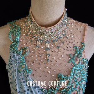 May include: A close-up of a light blue and teal dance leotard with a high neckline and a crystal embellishment design. The leotard is decorated with rhinestones and sequins. The text "Costume Couture by Naomi" is visible on the leotard.