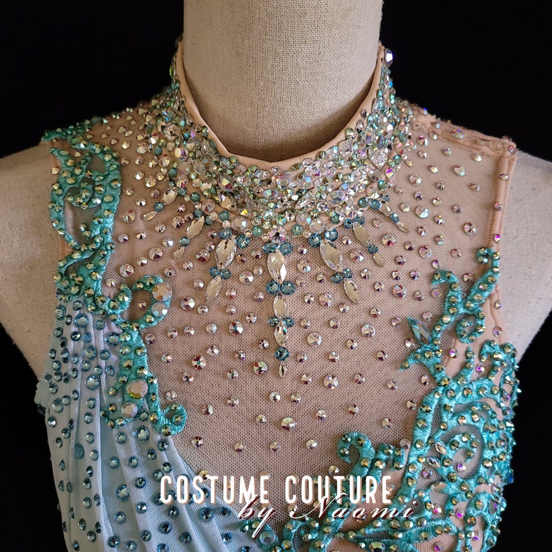 May include: A close-up of a light blue and teal dance leotard with a high neckline and a crystal embellishment design. The leotard is decorated with rhinestones and sequins. The text "Costume Couture by Naomi" is visible on the leotard.
