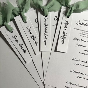 May include: A set of white place cards with black calligraphy names, including Joyce Anzola, Cesar Rincón, Luis Gabriel Rodriguez, and Marco Delgado. The cards are decorated with green ribbon.