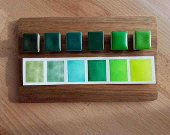May include: A set of eight watercolor paint pans in shades of green. The pans are arranged in two rows of four, with the top row showing the dry paint and the bottom row showing the wet paint.