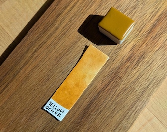 May include: A square, yellow ochre watercolor paint block and a swatch on a wooden surface. The swatch has the words "YELLOW OCHER" printed on it. The paint block is a square with a white base.