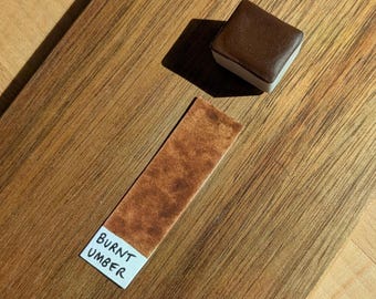 May include: A dark brown watercolour paint pan with a white base is on a wooden surface. Beside it is a colour swatch of the same shade, labelled "BURNT UMBER" on a white label.