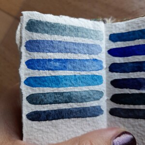 May include: A small, white, paper notebook with two pages open, showing a color chart of blue and indigo watercolor swatches.