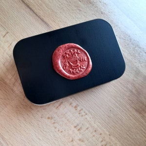 May include: A black metal tin with a red wax seal. The seal has a design of a plant with a pot and the word "Mom" written on it.