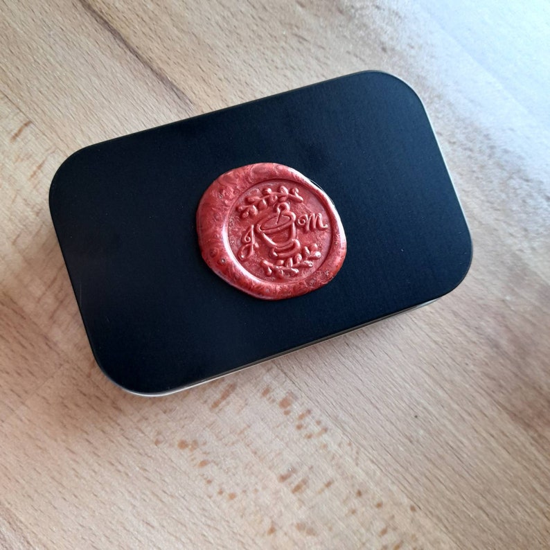 May include: A black metal tin with a red wax seal. The seal has a design of a plant with a pot and the word "Mom" written on it.