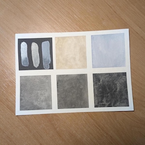 Puede incluir: Una tarjeta de muestras de acuarela con seis cuadrados y tres pinceladas verticales. Los cuadrados muestran varios tonos de gris, beige y azul. Las pinceladas son plateadas y azules sobre un fondo negro. La tarjeta está sobre una superficie de madera.