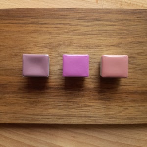 Puede incluir: Tres pequeñas bandejas cuadradas de pintura de acuarela. Las bandejas están dispuestas en una fila de izquierda a derecha. Los colores son un morado apagado, un rosa brillante y un melocotón claro.