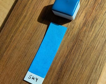 May include: A blue watercolor paint cube with a white base sits next to a blue color swatch labeled "SKY" on a wooden surface. The swatch is a rectangular strip of blue paper with the word "SKY" printed in black.