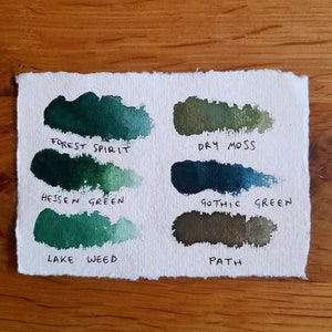 May include: A white paper swatch card with six different shades of green paint. The paint names are "Forest Spirit", "Dry Moss", "Hessen Green", "Gothic Green", "Lake Weed", and "Path".