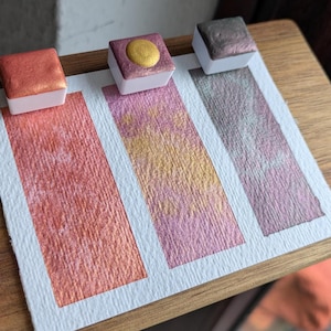 Op de afbeelding: Drie metallic aquarelverfmonsters op wit papier. De kleuren zijn koper-oranje, roze met gouden glans en grijs-paars. Elk verfmonster zit in een kleine witte vierkante container.