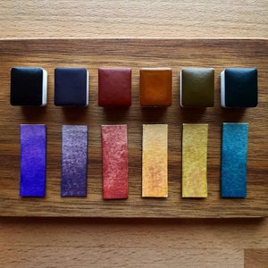 Könnte beinhalten: Satz mit acht Aquarellfarben in verschiedenen Farben, darunter Schwarz, Lila, Rot, Orange, Gelb, Grün und Blau. Die Farbtöpfe sind in einer Reihe auf einer Holzoberfläche angeordnet. Jeder Topf hat einen entsprechenden Farbmuster, der auf einem separaten Blatt Papier gemalt ist.