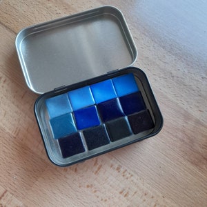May include: A metal tin with a lid open, revealing a set of 12 watercolor paint pans. The pans are arranged in a grid pattern and are filled with shades of blue and black.