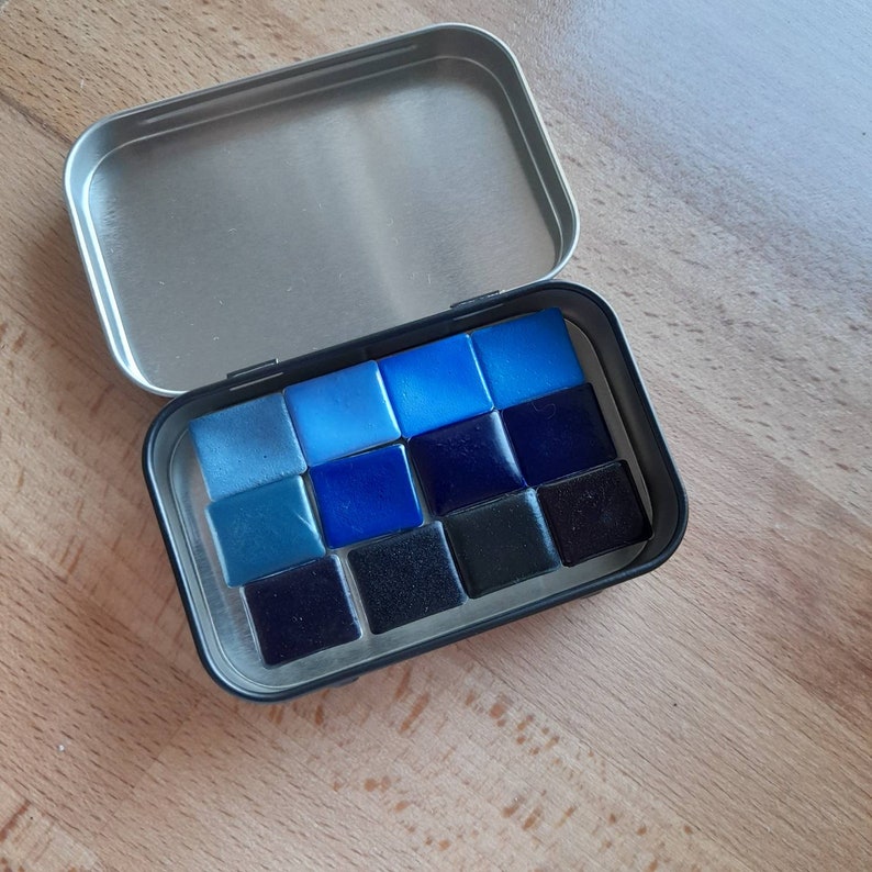 May include: A metal tin with a lid open, revealing a set of 12 watercolor paint pans. The pans are arranged in a grid pattern and are filled with shades of blue and black.