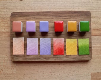 May include: A wooden board with eight small watercolor paint pans and eight corresponding color swatches. The pans are arranged in a row from left to right: peach, lavender, purple, red, yellow, green. The swatches are arranged in the same order below the pans.